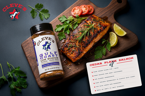 Grilled cedar plank salmon fillet topped with rosemary on a wood board, beside lemon, tomato, and a bottle of Cleve's Gulf Coast seafood seasoning.