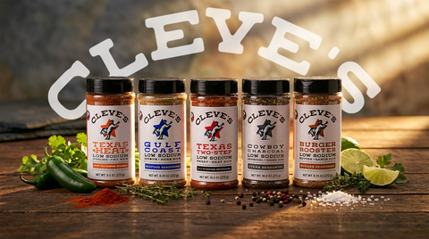 Cleve's low-sodium gourmet seasonings lineup: Texas Heat, Gulf Coast, Texas Two-Step, Cowboy Charcoal, and Burger Booster blends arranged on a rustic wood table with fresh jalapeños, lime, herbs, and peppercorns.